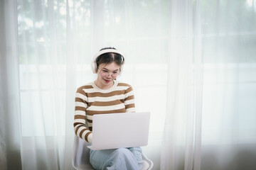 A person, who appears to be an East Asian female, wearing headphones and glasses, is casually using a laptop by a window.