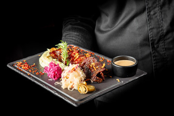 A chef holds a black plate with a gourmet dish featuring grilled meat garnished with rosemary, mashed potatoes, pickled vegetables, jalapenos, and a small bowl of sauce. Elegant presentation