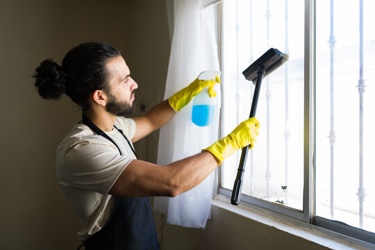 Young man cleaning windows at home wearing apron and gloves