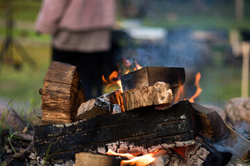 Detailed view of burning firewood in a campfire, with bright flames, smoke, and charred wood, set in a natural outdoor environment with a blurred figure in background.