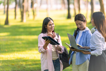 Group of Asian college students standing on green grass and discussing with laptop and books. Concept of teamwork, education, and outdoor learning in a natural environment.