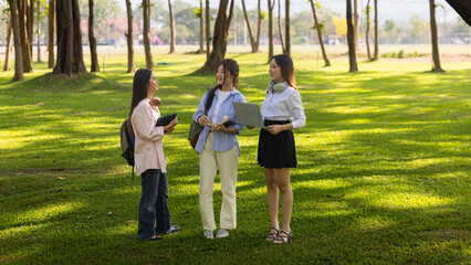 Group of Asian college students standing on green grass and discussing with laptop and books. Concept of teamwork, education, and outdoor learning in a natural environment.