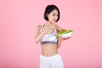 Fit Asian woman eating fresh vegetable salad with a wooden fork. Concept of healthy lifestyle, diet, and nutrition on pink background.
