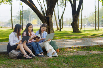 Three Asian university students sitting outdoors on campus using laptop, tablet, and books. Concept of teamwork, education, friendship, and digital learning in a natural setting.