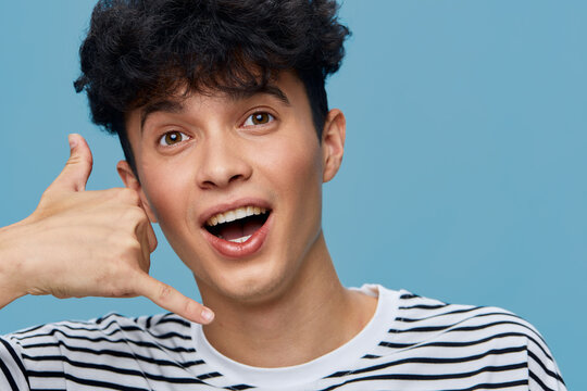 smiling man with curly hair making call me gesture wearing striped shirt on blue background, expressing friendly and playful emotion indoors.