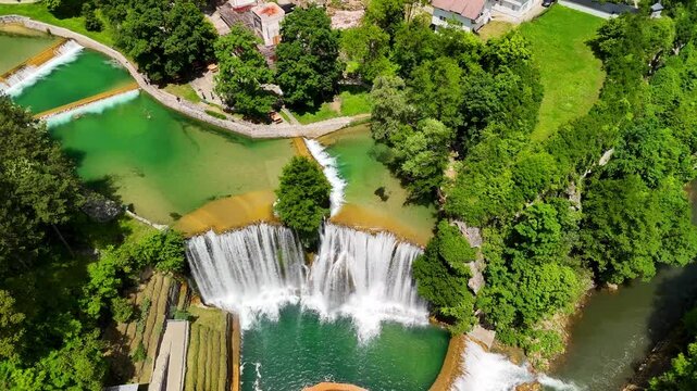 Aerial drone view of a picturesque waterfall cascading into a clear pool, surrounded by lush greenery and trees in a natural landscape. The Pliva Waterfall in the town of Jajce, Bosnia and Herzegovina