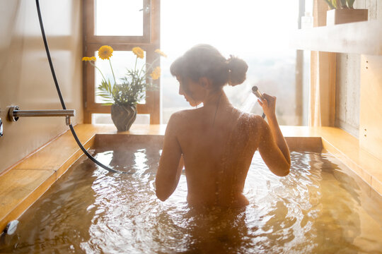 A woman enjoys a relaxing hot bath in a wooden tub with a panoramic view. Steam rises around her as she showers, surrounded by flowers and soft natural light