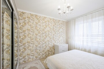 cozy bedroom features floral wallpaper, a white bed with a textured quilt, sheer curtains, and a chandelier. A sliding-door closet stands against the wall
