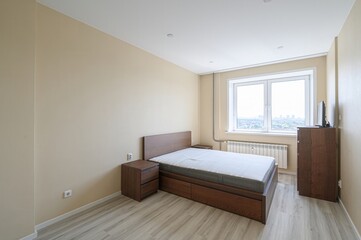 bedroom features a wooden bed, bedside table, and dresser. The walls are beige, and a window lets...