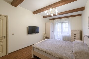 Cozy bedroom with a bed, TV, and wooden beams on the ceiling. A window with sheer curtains lets in natural light.