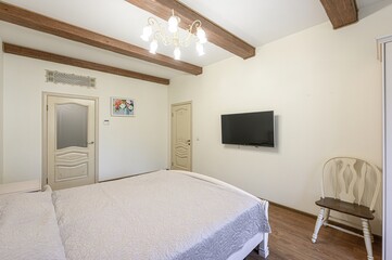 Cozy bedroom interior with a bed, TV, chair, and wooden beams on the ceiling. Two doors and a small picture are also visible