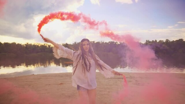 Young adult happy woman dancing spinning hands holding smoke bomb red color. river bank enjoy life blue sky, black long hair boho style hippie girl back rear view. feather on head. fun woman summer