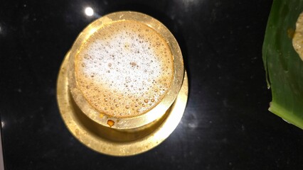 Glass of steaming hot tasty filter coffee in brass tumbler of glass and bowl. Hot south Indian filter coffee with froth and cream.