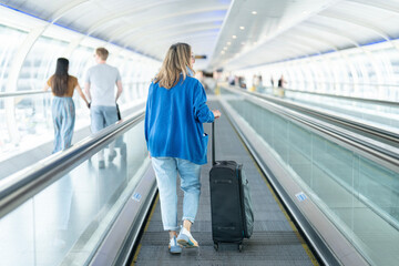 Woman traveling through a modern airport terminal with a suitcase on a moving walkway during...