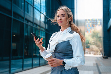Professional woman in business attire checks phone while holding coffee in urban setting during the...