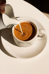 Minimalistic top-angle photo of a rustic white ceramic cup on a matching saucer, as milk is being poured into freshly brewed coffee. The swirl is clearly visible at the center of the cup