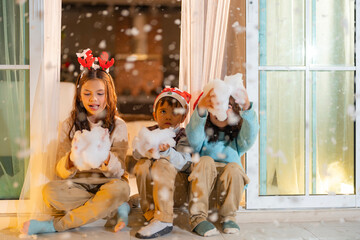 Three children wearing Christmas accessories sit at the doorway holding artificial snow, enjoying the holiday spirit with joy and curiosity in a cozy indoor winter-themed setting.
