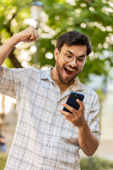 Indian young man using mobile smartphone celebrating win good message news, lottery casino jackpot victory, giveaway online outdoors. Hispanic guy on urban city street. Town lifestyles. Vertical.