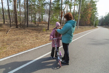 Little girl riding scooter while walking with her grandmother in the forest on a sunny day