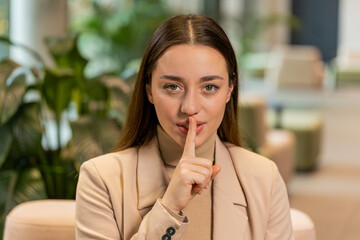 Shh be quiet please, keep my secret. Portrait of young Caucasian businesswoman presses index finger to lips makes silence gesture sign do not tell anything. Pretty woman girl sitting in office lobby.