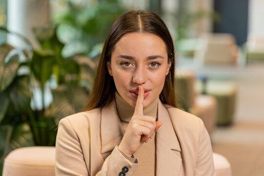 Shh be quiet please, keep my secret. Portrait of young Caucasian businesswoman presses index finger to lips makes silence gesture sign do not tell anything. Pretty woman girl sitting in office lobby.
