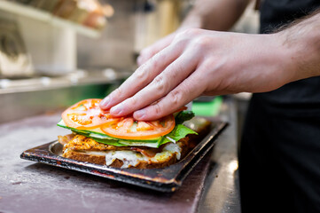 Close-up of hands preparing a sandwich with fresh ingredients—tomato slices, lettuce, and melted cheese—on a kitchen counter. The image captures the process of assembling a delicious homemade meal.