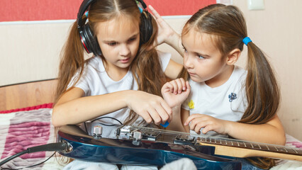 Two child girls are sitting on the bed with a electric guitar. The older sister teaches the younger...
