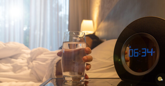 Early morning scene of a person in bed reaching for a glass of water beside a digital alarm clock showing 6:34 AM. Soft sunlight streams through sheer curtains in a cozy, peaceful bedroom setting.