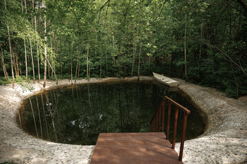  Rowboat rests beside a tranquil forest pond surrounded by lush greenery and pebbled shore,...