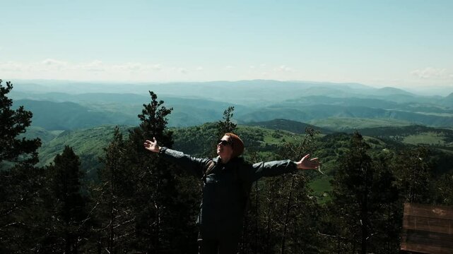 Young woman hiker with backpack and orange hat stretches her arms wide while enjoying a panoramic mountain view from Tornik peak in Zlatibor, Serbia during spring. Solo travel concept