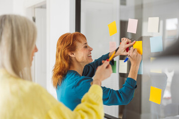 Businesswomen planning strategy using sticky notes on glass wall in office