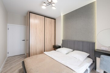 modern bedroom featuring a bed with white linens, a gray headboard, and a beige comforter. A wooden closet, bedside table, and a vanity with a round mirror complete the space