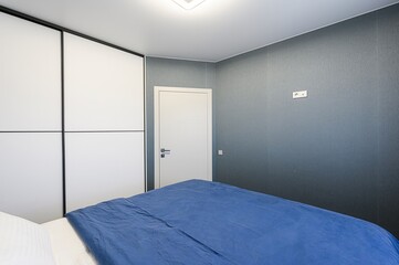 modern bedroom interior featuring a blue bedspread, white closet, gray wall, and a white door. The room has a minimalist design