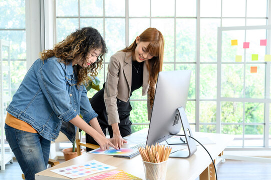 Happy two young female graphic designers discussing with colleague in office. Creative people working together using computer at desk.