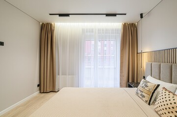 bright bedroom scene featuring a neatly made bed, light beige walls, wood flooring, and window with sheer and gold curtains