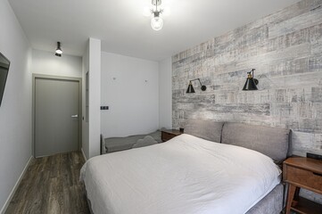 minimalist bedroom with a bed against a faux wood wall, flanked by nightstands and sconces. A small...