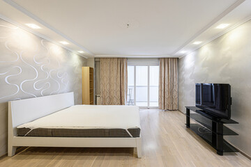 Bright bedroom with a white bed, a black TV stand, and curtains around a sliding glass door....