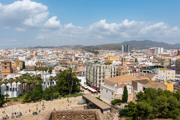 Elevated view from Alcazaba of Málaga’s cathedral and white city rooftops, framed by palm trees and clouds over the mountains.