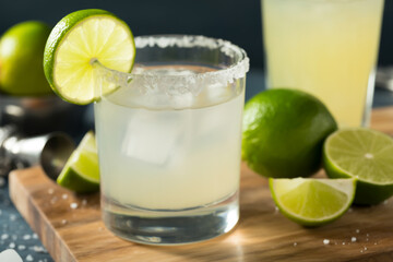 A margarita cocktail with lime garnish and salt rim on a wooden board with more limes nearby