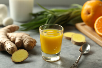 A glass of ginger shot with ginger root orange spoon and green herbs on a gray surface in soft light