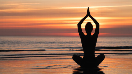 Silhouette of person meditating on beach at sunset with hands in prayer position overhead for health and wellness