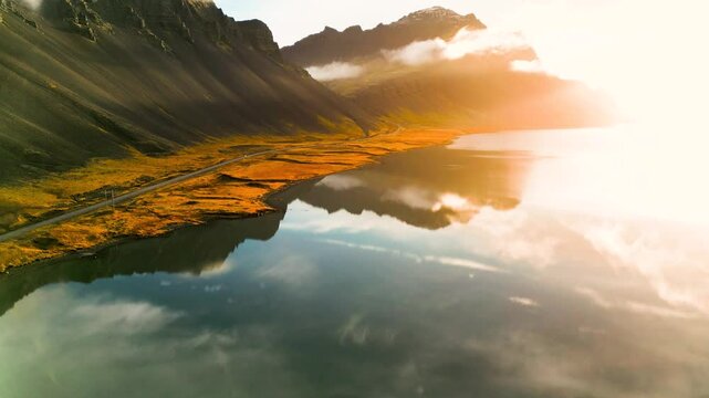 Vulkanische K&uuml;stenlandschaft in Island im goldenen Morgenlicht mit ruhiger Wasserfl&auml;che, Spiegelung der Berge und einer schmalen Stra&szlig;e entlang des Ufers in herbstlicher Umgebung