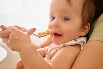 A close-up shot captures a baby with bright blue eyes, with a bib on, opening its mouth to be fed a spoonful of food by an unseen hand.