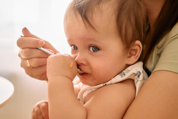 A close-up captures a baby with wide blue eyes, being fed a spoonful of food, while holding the...