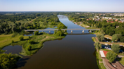 Scenic aerial view of a riverside town with a historic castle, marina, pedestrian bridges, and lush greenery on a sunny summer day. Calm river, clear sky, and charming European architecture.