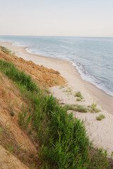 A coastal landscape features a tall, sandy cliff covered with patches of green vegetation, sloping...
