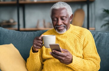 an elderly Black man sitting on a couch, holding a coffee mug and using a smartphone at home, wearing a yellow sweater