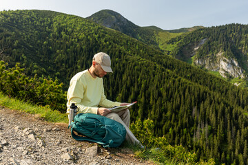 man studying map sitting on edge of cliff overlooking mountains. hiking mountains