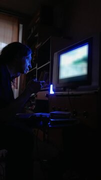 An intensely concentrated and immersive mood is captured as a man with long hair plays a video game on an older television in a dimly lit room, his face illuminated by the screen's glow
