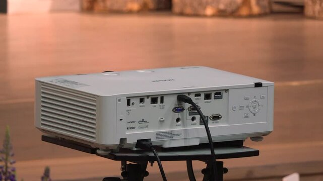 Close-up of a white Epson projector showing rear ports and cables, placed on a tripod stand in a wooden-floored indoor space.
